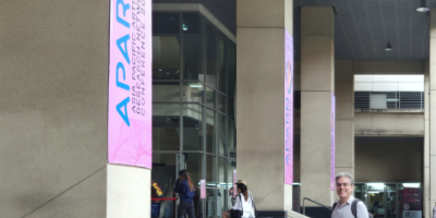 Modern conference venue entrance featuring prominent pink APARN banners with blue lettering displayed on concrete pillars. The contemporary building showcases floor-to-ceiling glass windows and sleek architectural design. Michail Semoglou, wearing glasses and a white shirt, stands in the foreground among other conference attendees at the APARN 2025 conference venue in Bangkok, Thailand, where he presented his research on Algorithmic Interpretations of Epirotic Silverwork.