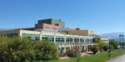 Modern university library building with a multi-colored facade featuring red brick, beige stone, and green architectural elements against a clear blue sky. The three-story structure showcases large glass windows and a contemporary design, with landscaped greenery in the foreground and mountains visible in the background.